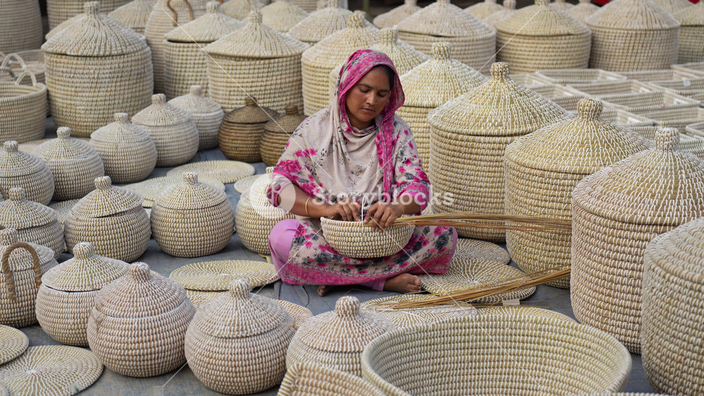 Bogura, Bangladesh - 14 November 2023: View of a woman sitting on the ground and weaving traditional handmade baskets surrounded by finished products.