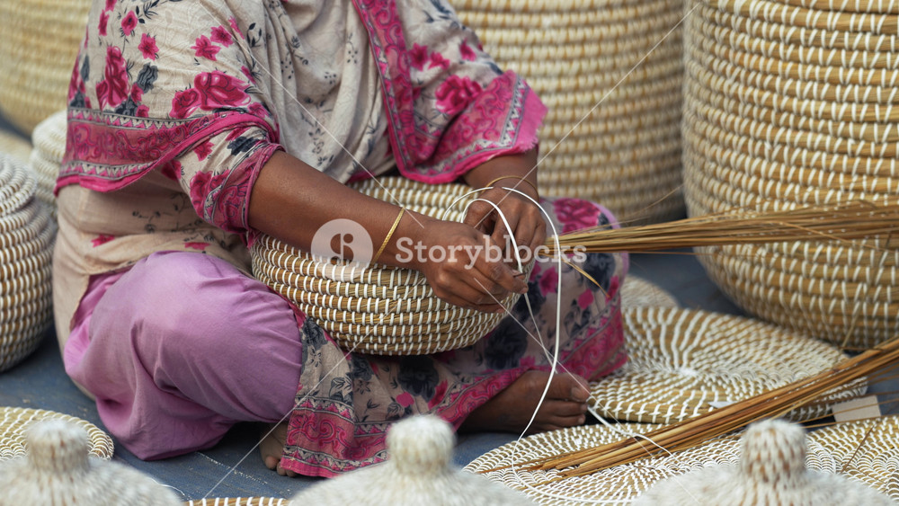 Bogura, Bangladesh - 14 November 2023: View of a woman sitting on the ground and weaving traditional baskets with natural fibers surrounded by finished products.