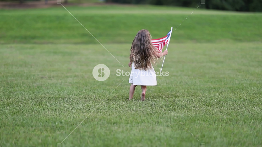A cheerful girl runs through a green park holding a small American flag. Her playful movement captures the spirit of childhood freedom and a