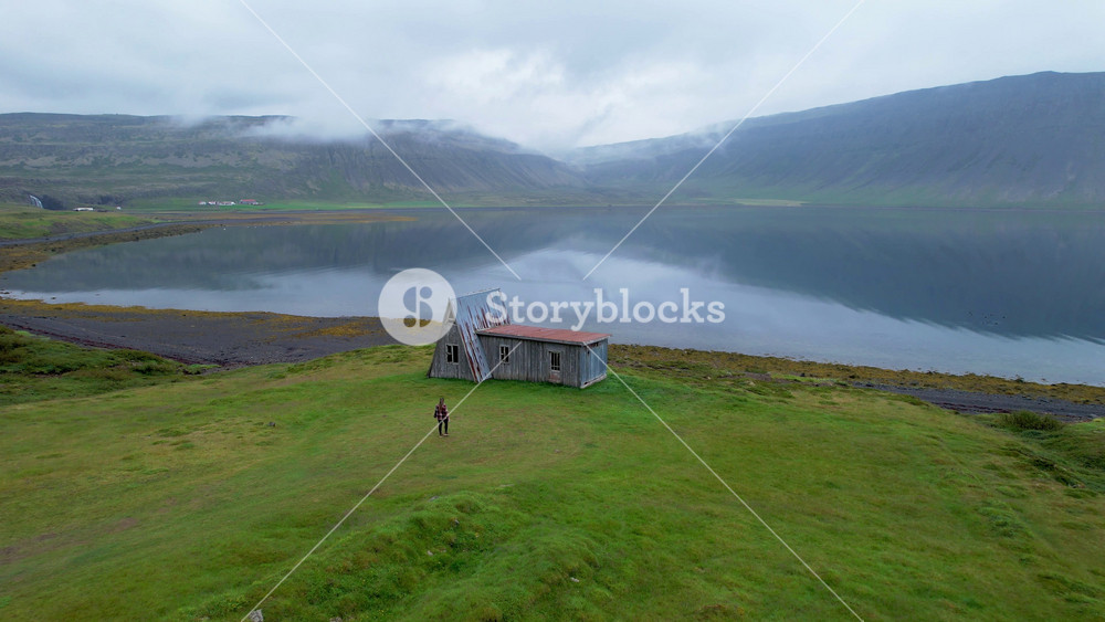 Travel Girl Walks Toward A Frame Cabin in West Fjords Iceland Drone From Side Towards Her. Caucasian Female Tourist Explores Iceland Cabin in Remote Landscape by Fjord Coast..