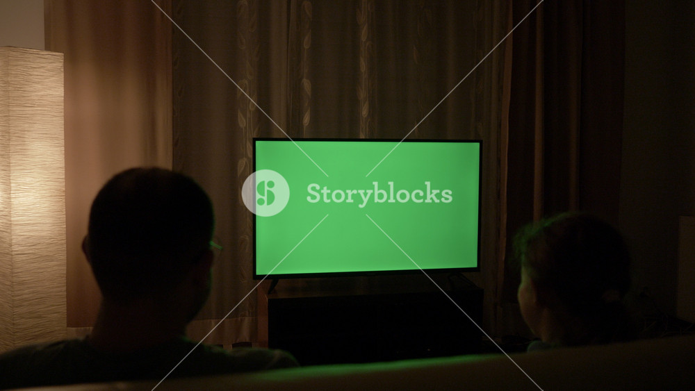 Family watching television with blank green chroma key screen in dark living room. People watch TV with green chroma key screen. Family Sitting Together Sofa In Their Living Room Night Watching TV.