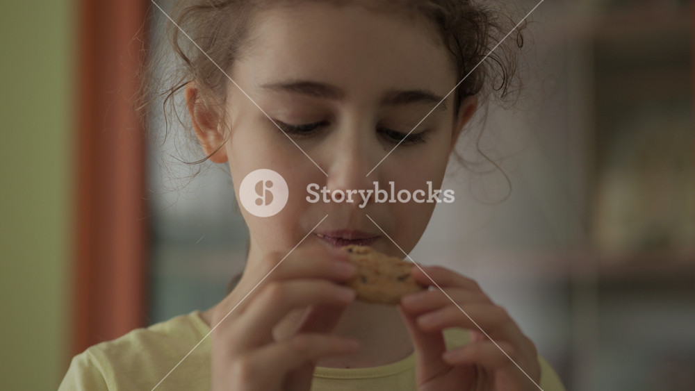 Little Girl Eating a Cookie. Looking at Camera and Smiling. Cute Child Hungry Eating Cookie Enjoying Delicious Treat at Home in Kitchen. Junk Food, Unhealthy Nutrition Sugar.