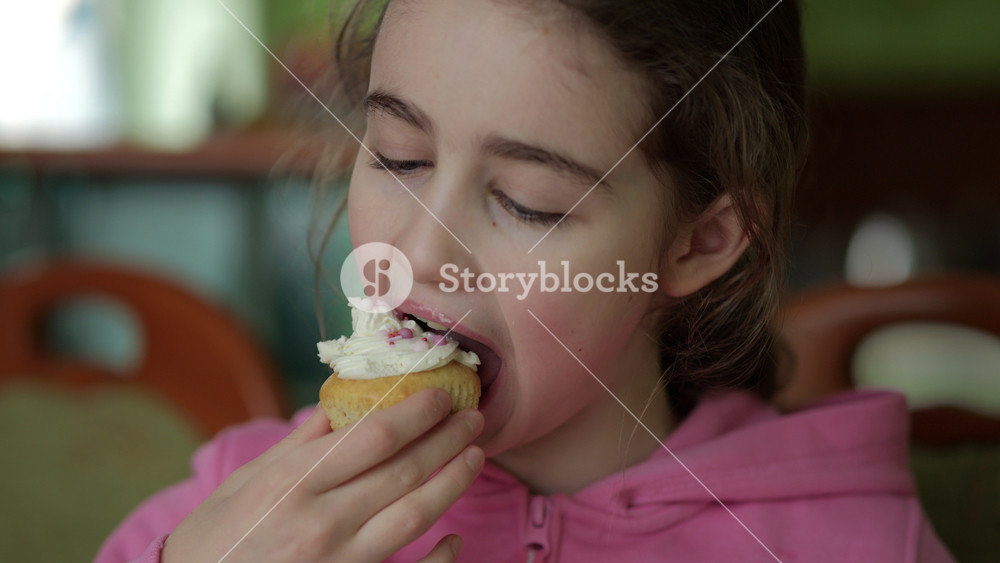 Child Eating Muffins Cupcakes Decorated With Sugar Cream. Girl Enjoying Vanilla Cupcakes with Frosting and Sprinkles. Unhealthy Junk Food. Hungry Sweet Tooth Child Bites Muffin with Sugar Frosting.