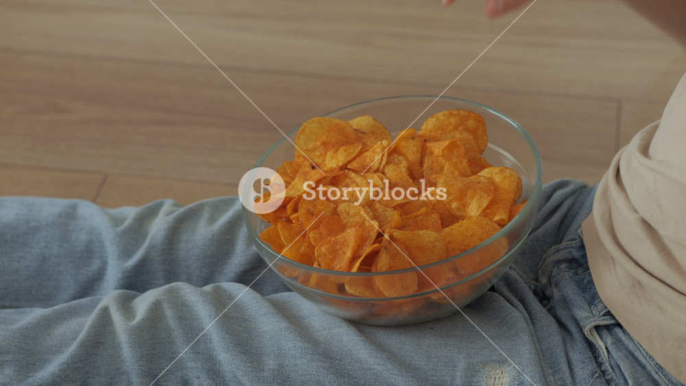 Girl Eating Chips From Bowl While Sitting on Floor in Living Room. Female Eating Unhealthy Potatoes Crisps. Snack on Junk Food. Child Eats Fast Food, Junk Food at Home Watching TV. Childhood Obesity.