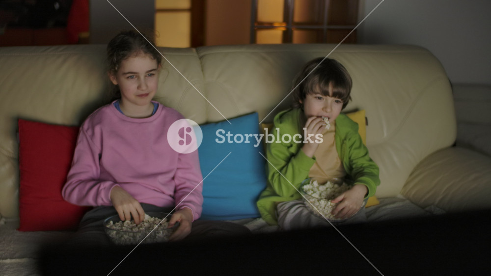 Children Sitting On Sofa In Living Room Evening Eating Popcorn and Watching TV Together on Weekend in Apartment. Brother and Sister Watching Movie at Home in Living Room at Night. Kids Entertainment.