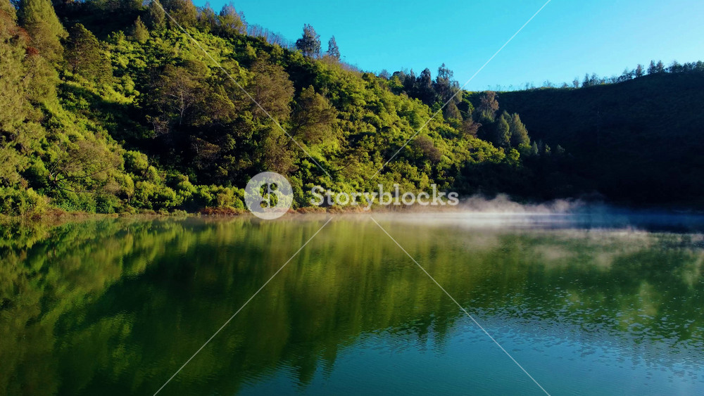 Experience the ethereal beauty of Ranu Pani Lake at the foot of Mount Semeru, where morning mist dances above the tranquil waters. Surrounded by lush green trees, this serene scene captures nature's
