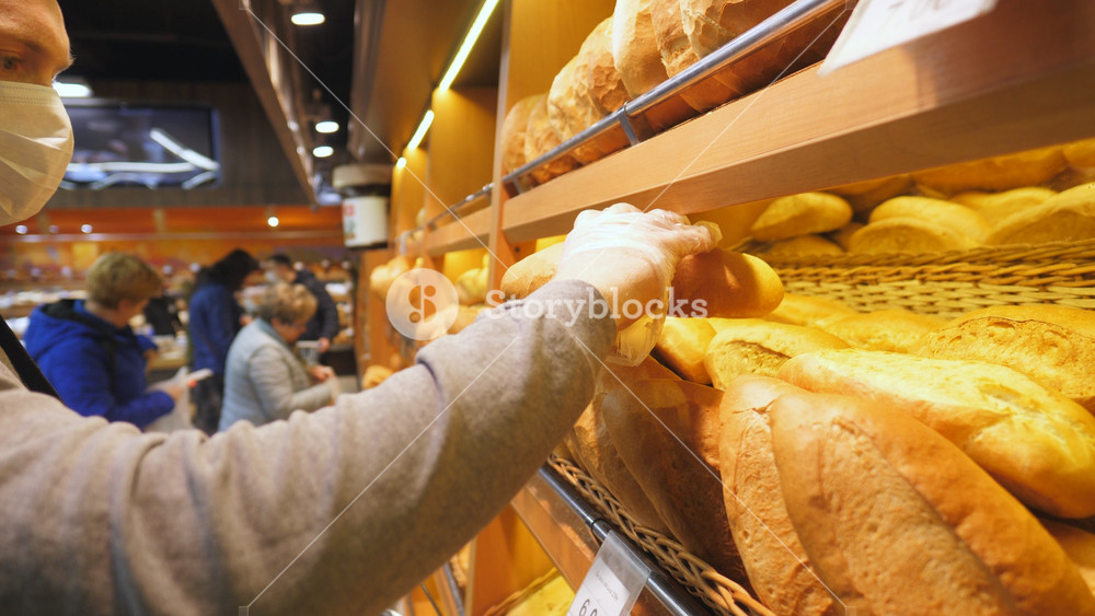 Young man with medical face mask chooses a fresh bread loaf in the supermarket. Guy selecting food at produce department of shop at quarantine time. Shopping in the grocery during coronavirus pandemic.