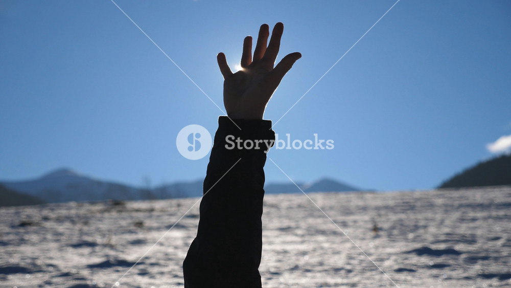 Young man lies down on snowy field and his arm reaches to sun. Bright sunlight breaks through fingers of outstretched male hand to blue sky. Concept of winter vacation or holiday. Close up Slow motion.