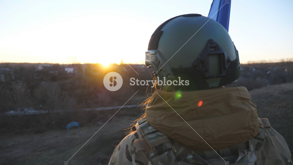Ukrainian army woman stands to the peak of hill with waving blue-yellow banner at sunset. Young female soldier in camouflage uniform lifting flag of Ukraine. Resistance to russian invasion. Crane shot.