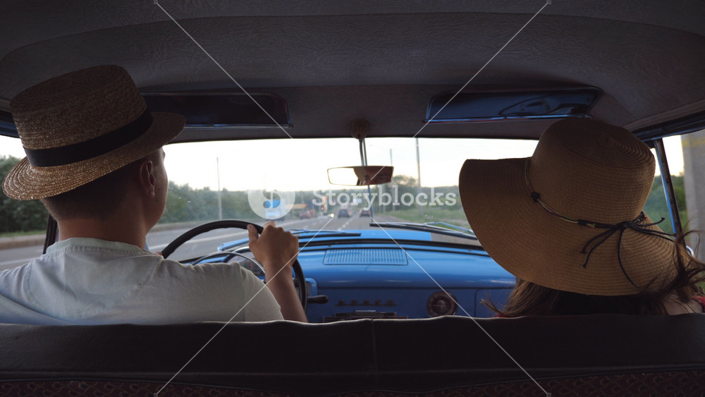 Young couple in hats riding at vintage car during road trip. Unrecognizable man and woman sitting at the front seat of old retro car. Summer travel concept. Slow motion Rear back view Close up.