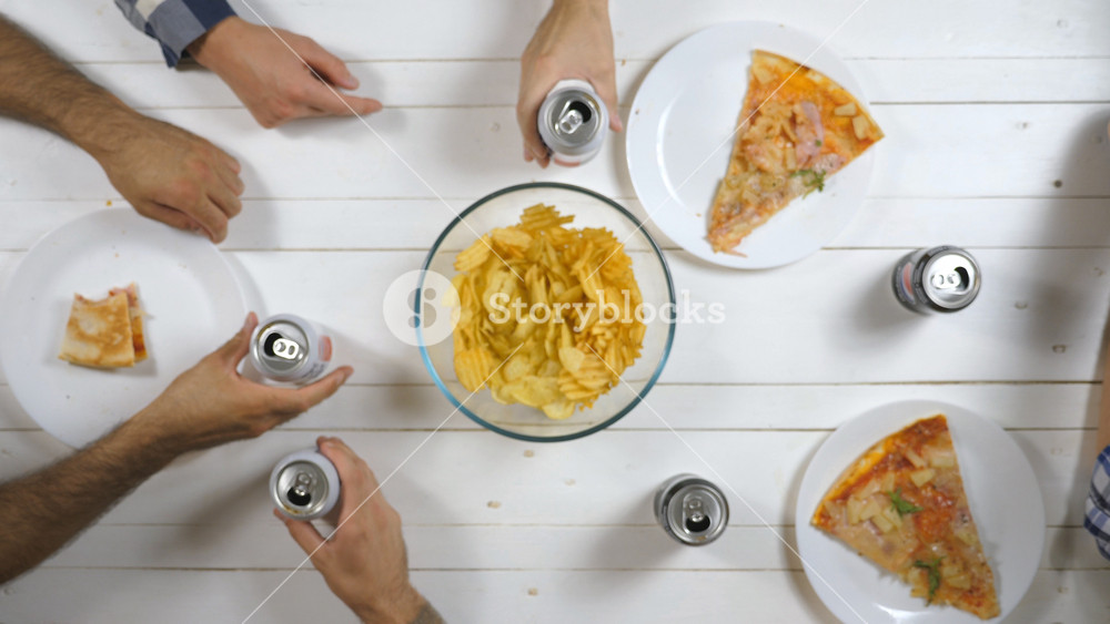 Top view group of best friends sitting at the table and clicking aluminum cans with soda on wooden table background. Male and female hands drinking cola and eating snacks indoor. Slow motion
