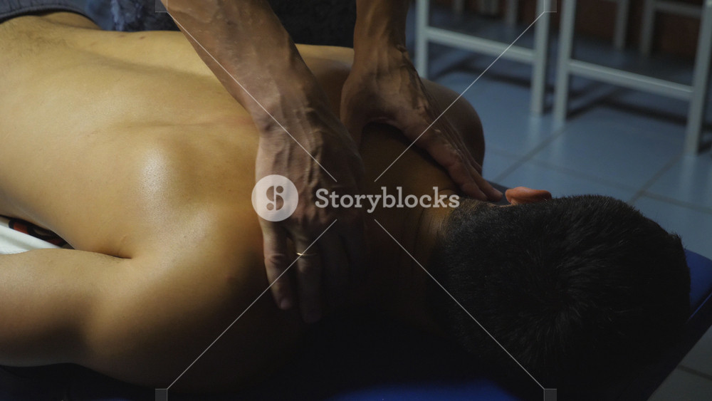 Close up to hands of masseur massaging shoulders of sportsman in salon. Male arms of professional massagist doing healing rubdown of back to young athlete in parlor. Concept of healthy lifestyle.