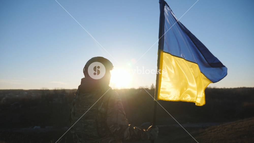Ukrainian army girl stands with lifted national banner against background of sunset. Young female soldier in military uniform holds a waving flag of Ukraine to peak of hill. Victory of war. Dolly shot.