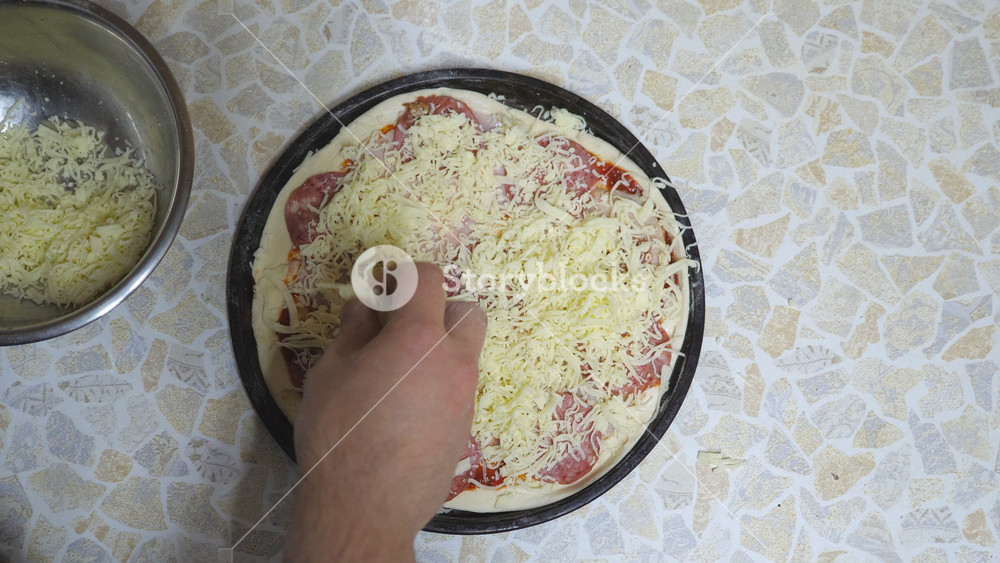 POV male hands of cook putting grated cheese on pastry with ingredients in metal form at cuisine. Chef making a pepperoni pizza on wooden surface at kitchen. Food preparing concept. Top view Slow mo