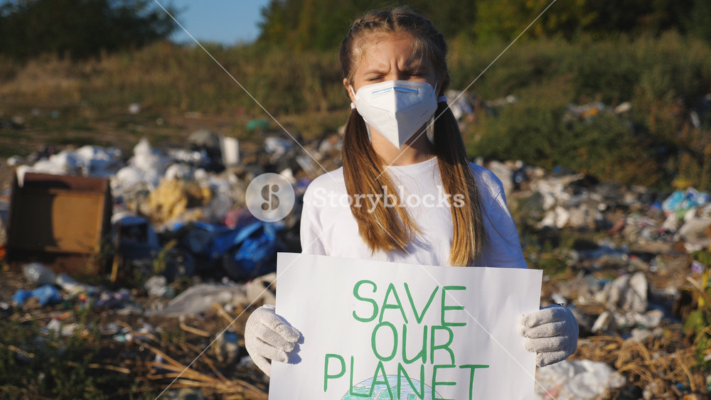 Portrait of small girl holds in hands poster of environmental movement for saving planet. Little female eco activist in protective mask looking into camera against the background of landfill. Close up.