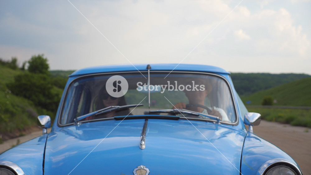 Young couple in hats riding in vintage car at summer travel. Man and woman sitting at the front seat of old retro car during ride. Concept of road trip at holidays . Slow motion Close up Front view.