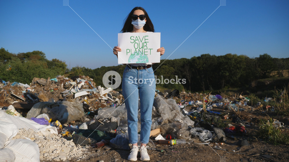 Young girl in medical protective mask looking into camera and standing against the background of landfill. Female eco activist holding in hands placard of environmental movement for saving planet.