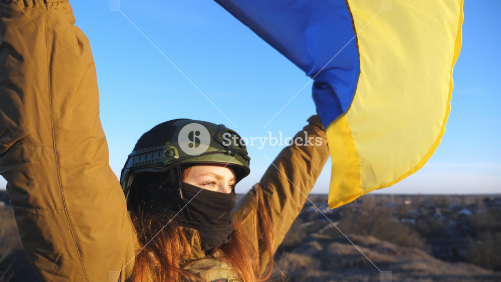 Female soldier of ukrainian army holding waving flag of Ukraine. Girl in military uniform and helmet lifted up yellow-blue flag. Victory against russian aggression. Invasion resistance concept. Slowmo.