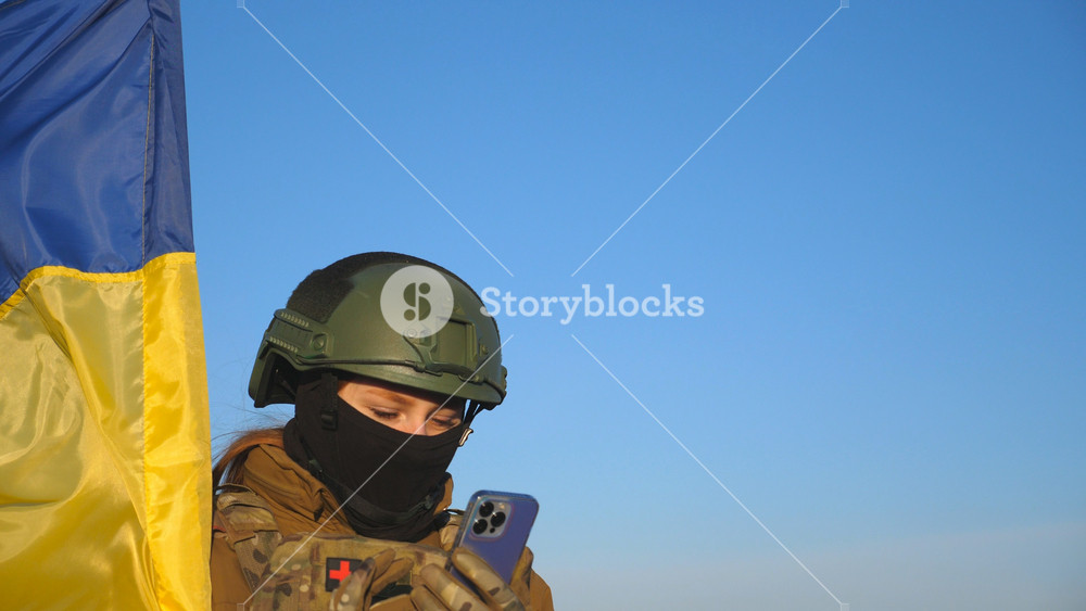 Female ukrainian army soldier using smartphone to read news or messaging with family. Girl in military uniform and helmet holding waving flag of Ukraine. Victory against russian aggression. Close up.