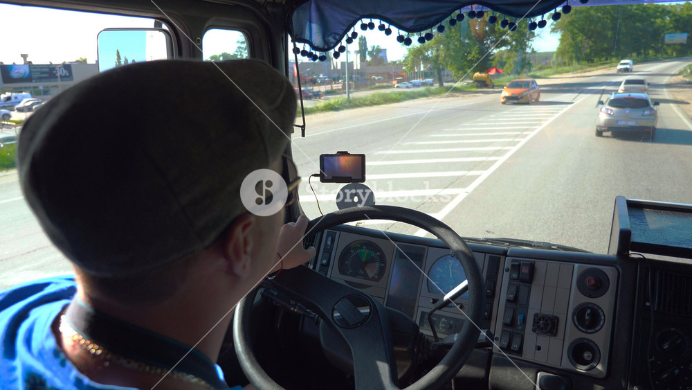 Unrecognizable lorry driver riding on city road at sunny day. Man in cap and sunglasses controlling his truck attentive watching road. View of traffic from inside cabin. Inside shot Slow mo Close up