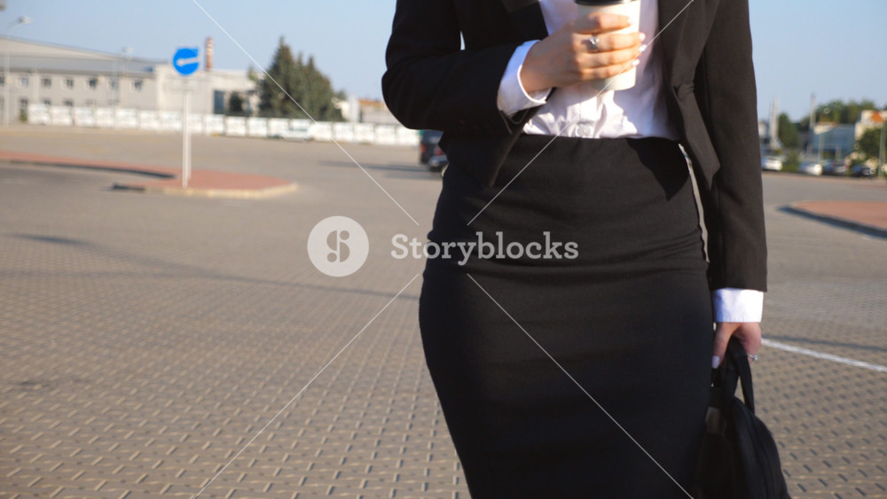 Young businesswoman with cup of coffee walking near auto parking at sunny day. Female legs in high heels shoes stepping in urban street