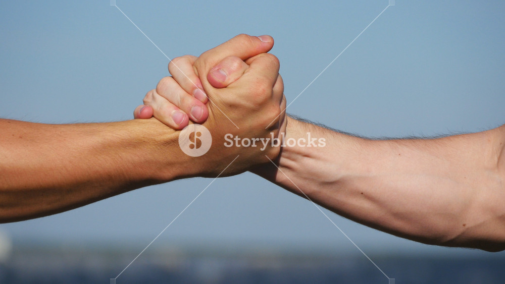Friendly handshake of two unrecognizable muscular white men on blue sky background. Shaking of male arms outdoor. Two strong men having firm handshake outside. Teamwork and friendship. Close-up.