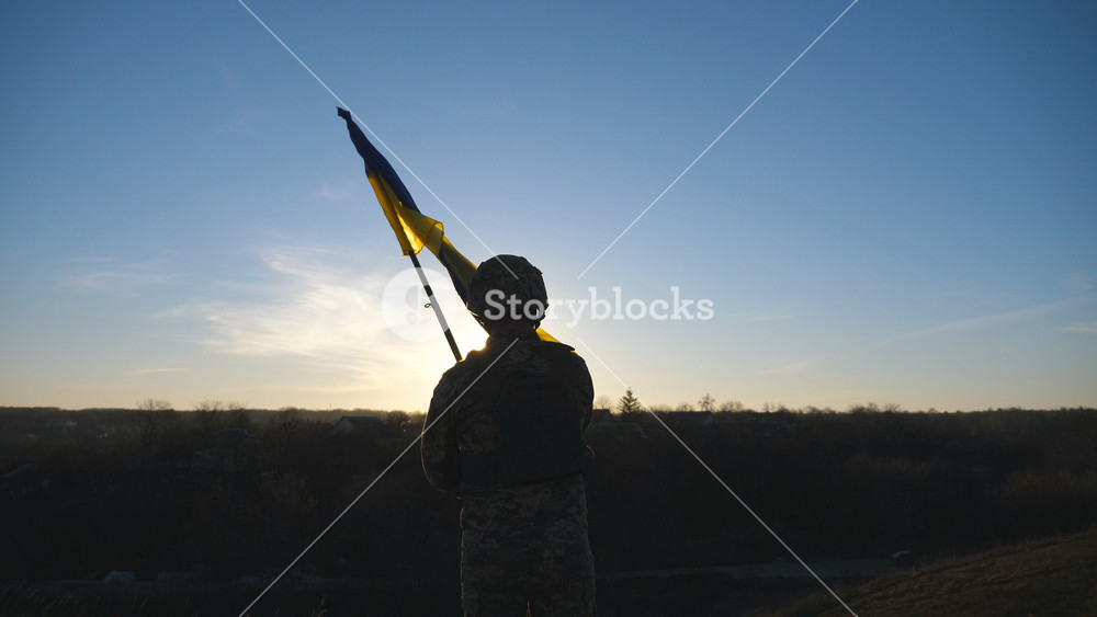 Soldier of ukrainian army stands at hill and holds waving flag of Ukraine. Man in military uniform and helmet lifted up flag against sunset. Victory against russian aggression. Invasion resistance.