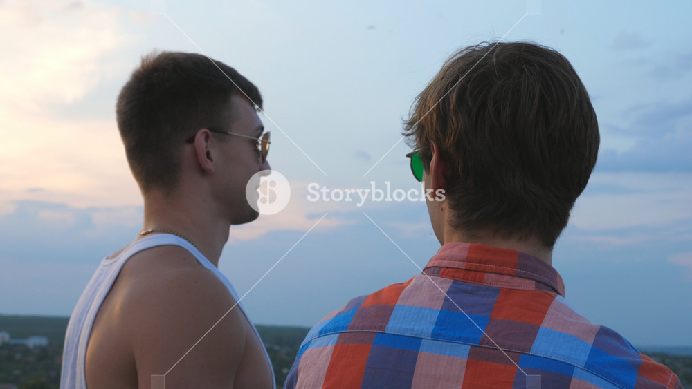Dolly shot of young male couple in sunglasses standing on the edge of rooftop and talking. Gay men relaxing on roof of high-rise building and enjoying beautiful cityscape at sunrise. Close up Slow motion.