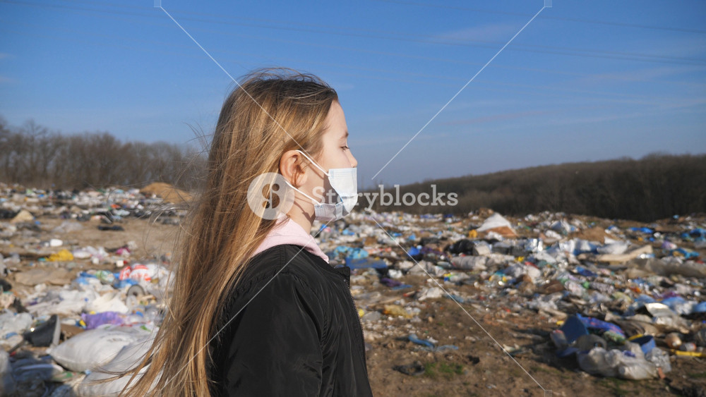 Small female child in medical protective mask goes along road against the background of garbage dump. Little girl with blonde hair walks near a lot of trash at countryside. Environmental pollution