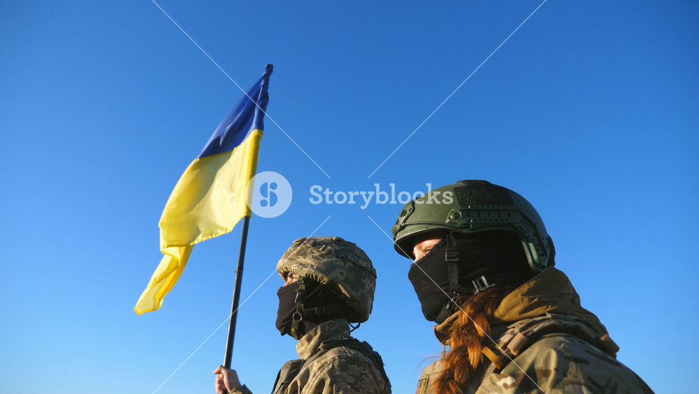 Young soldiers of ukrainian army standing at peak of hill with raised flag of Ukraine. Military couple in camouflage uniform taking each other hands as a symbol of support. Victory at war. Crane shot