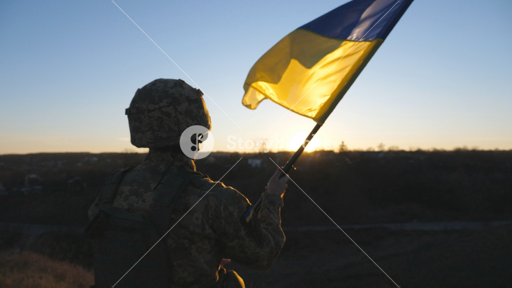Military man in uniform waving flag of Ukraine against background of sunset. Male ukrainian army soldier lifting national banner at hill in honor of the victory against russian aggression. Rear view.