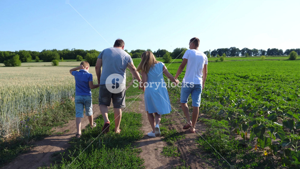 Young parents with two sons hold hands of each other walking along rural road near cultivated fields. Mother and father with children going along path near green meadows. Family spending time together.