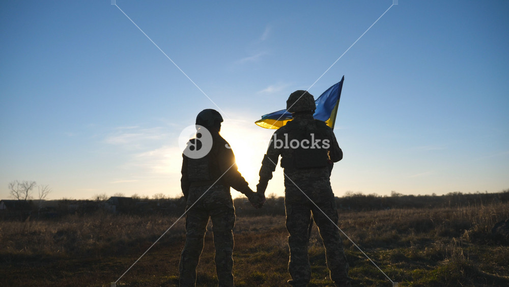 Ukrainian army soldiers stands outdoor at sunset time and waves flag of Ukraine. People in military uniform lifted up flag against blue sky