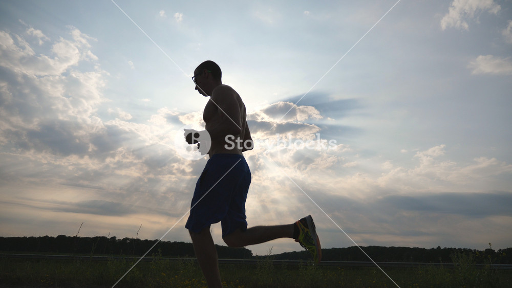 Silhouette of muscular man jogging in the country road at sunset. Male jogger training for marathon run outdoor. Athlete exercising and running against blue sky. Sport and active lifestyle Slow motion.