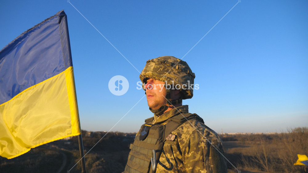 Ukrainian army soldier holding waving flag of Ukraine. Portrait of man in military uniform and helmet lifted up flag in hill. Victory against russian aggression. Invasion resistance concept. Slow mo.