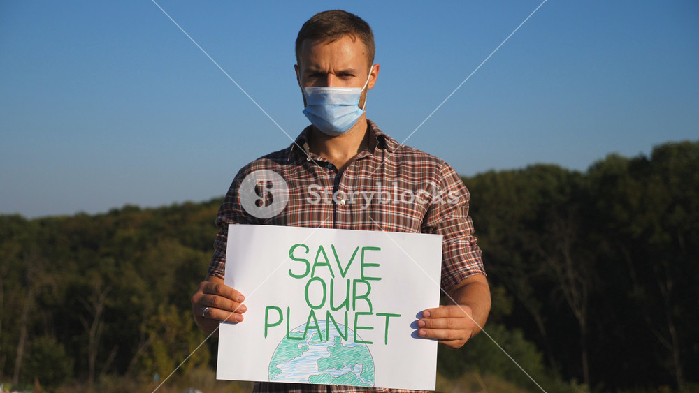 Portrait of young man holding in hands poster of environmental movement for saving planet. Male eco activist in protective mask looking into camera and advocating for nature conservation. Close up.