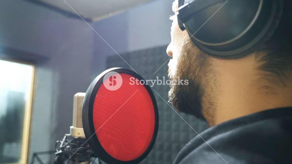 Male singer with beard in headphones singing song into the microphone at sound studio. Young man recording new melody or song. Working of creative musician. Show business concept. Slow motion.