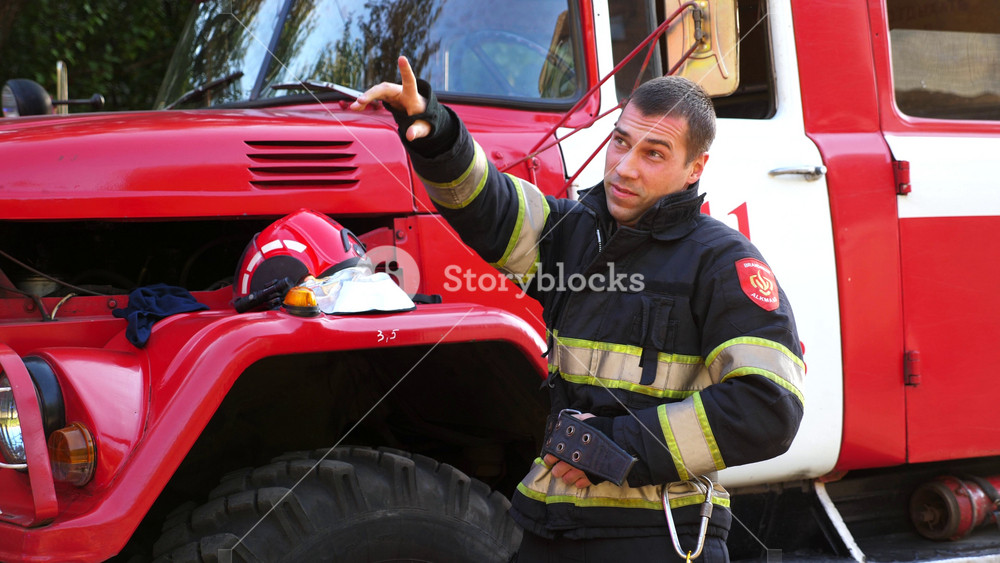 Young tired fireman unzipping protective workwear after hard work near a fire engine. Male firefighter after fire against background of a big red truck. Concept of saving lives and heroic profession.