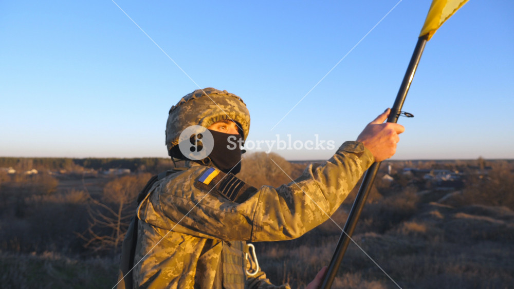 Dolly shot of male ukrainian army soldier lifting national banner in hill. Young man in military uniform waving flag of Ukraine against blue sky. Victory at war. Resistance to russian invasion concept.