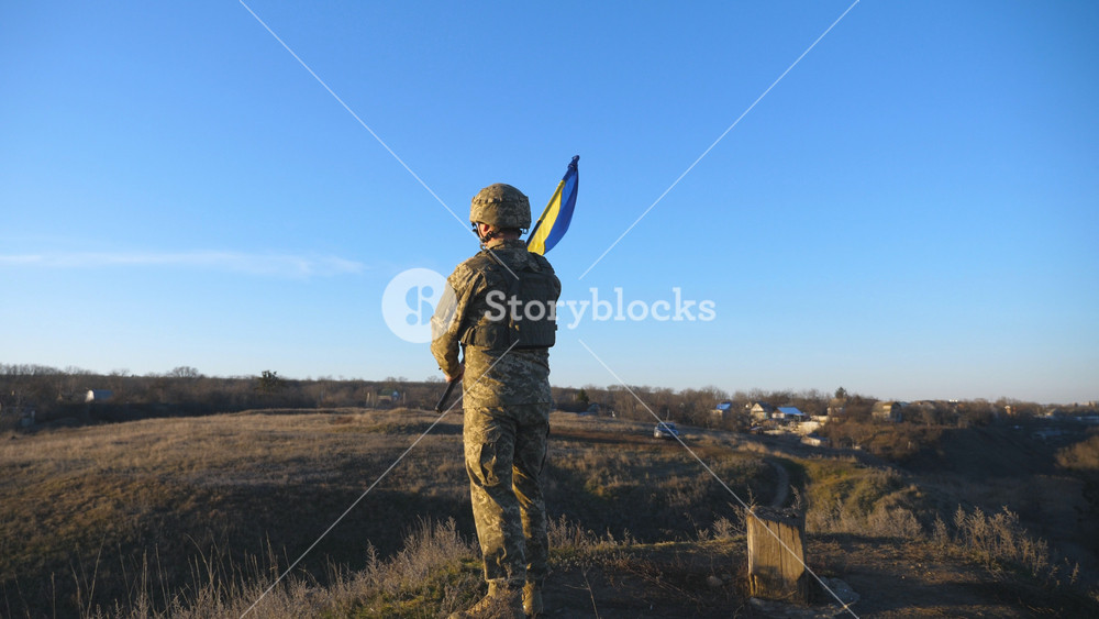 Portrait of soldier in military uniform lifted up flag of Ukraine in hill. Ukrainian army man holding waving flag. Victory against russian aggression. Invasion resistance concept. Slow motion.