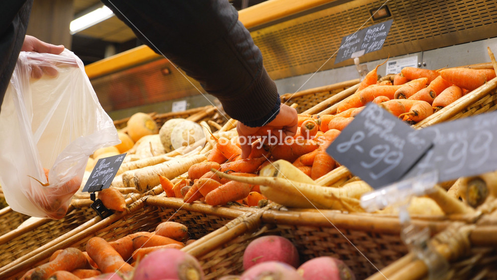 Hand of man selecting fresh carrot in grocery store or vegetable shop. Male buyer choosing vegetables and putting them in plastic bag. Variety of goods in supermarket. Shopping concept. Close up.