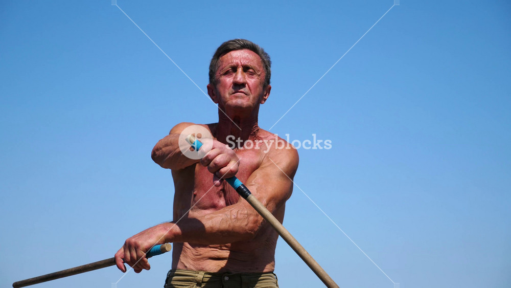 Older sportsman demonstrating wing chun kung fu against blue sea at background. Adult male athlete showing fighting techniques with wooden sticks at sunny summer day. Old man training martial arts.
