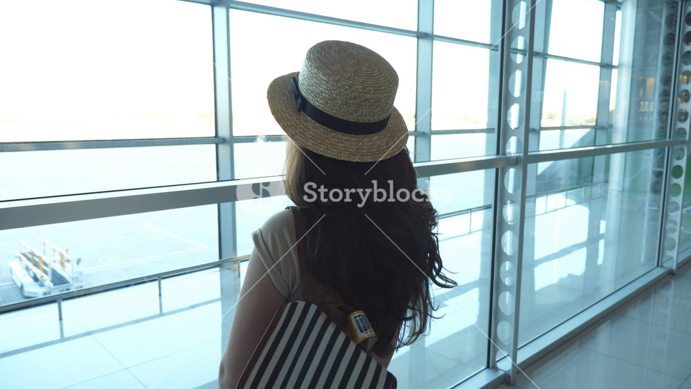 Young woman in hat with backpack standing in terminal of airport and looking at window. Girl waiting for her airplane. Tourism and travel concept. Rear back view Slow motion