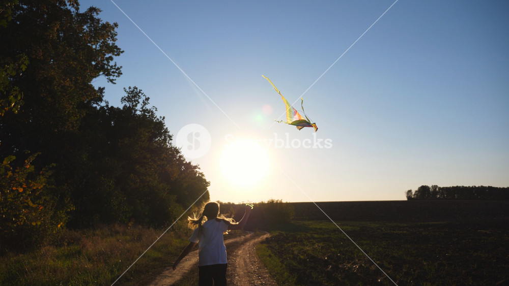 Follow to happy child jogging with rainbow kite on rural path with sunlight at background. Cute little girl plays with flying toy at countryside. Carefree kid having fun at nature and enjoying freedom