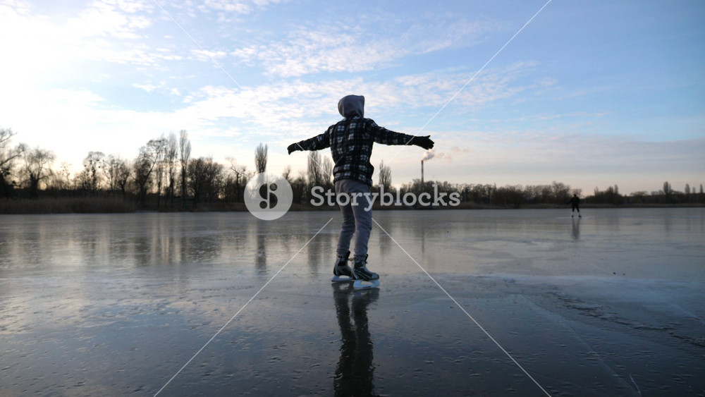 Following to young man shod in figure skates sliding on ice. Guy skating on frozen river. Sky reflected in icy surface of lake. Active leisure at cold winter. Concept of healthy lifestyle. Slow motion.
