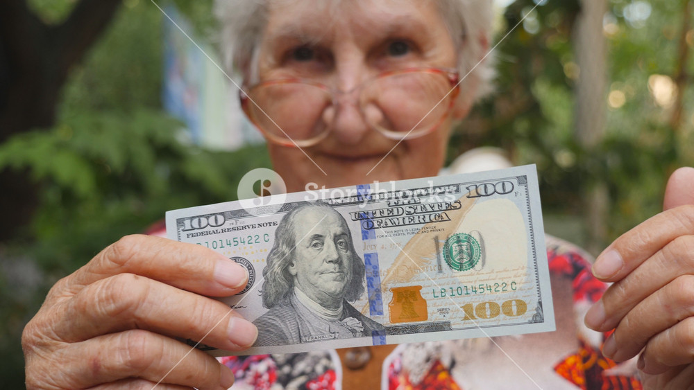 Old woman in eyeglasses showing one hundred dollar bill into camera outdoor. Happy grandmother holding foreign currency outside. Money