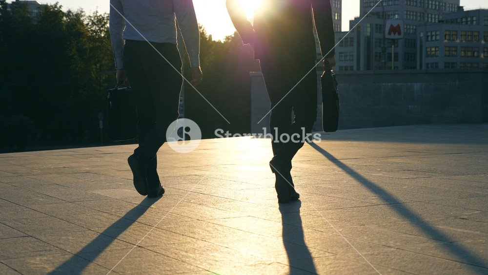 Two businessmen with briefcases walking in city street with sun flare at background. Business men commute to work together. Confident guys being on his way to office. Colleagues going outdoor. Slow mo.
