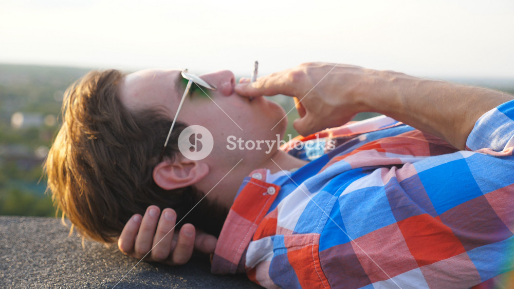 Handsome guy in sunglasses lying of the edge on rooftop and smoking cigarette with blurred cityscape background. Young man resting on roof of high-rise building and smiling at sunny day. Dolly shot.