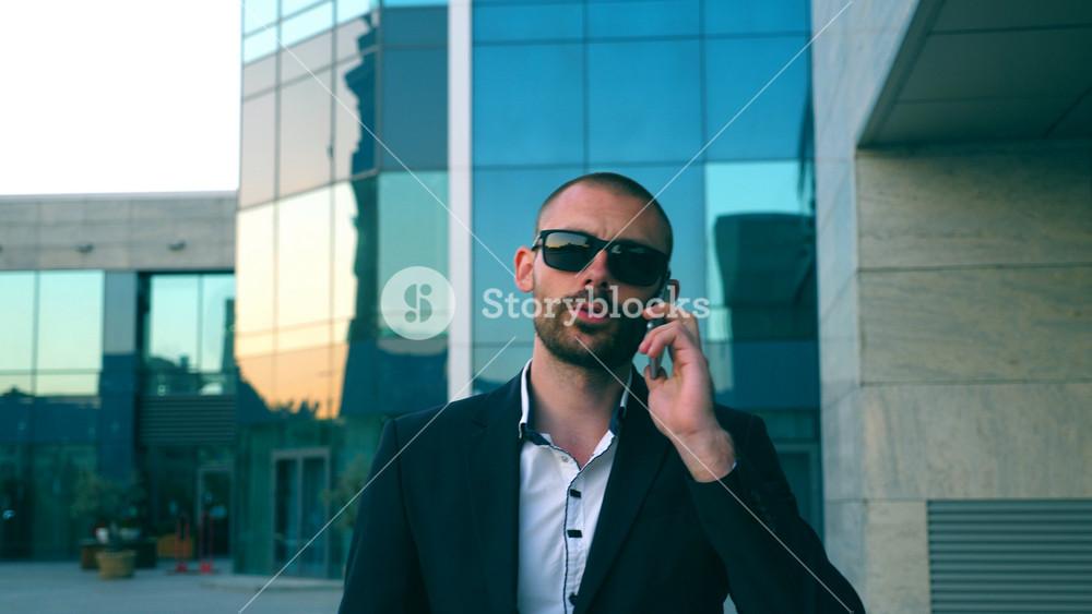 Portrait of businessman in sunglasses talking on phone and walking in street. Young man having business conversation during commuting to work. Confident guy in suit being on his way to office building.