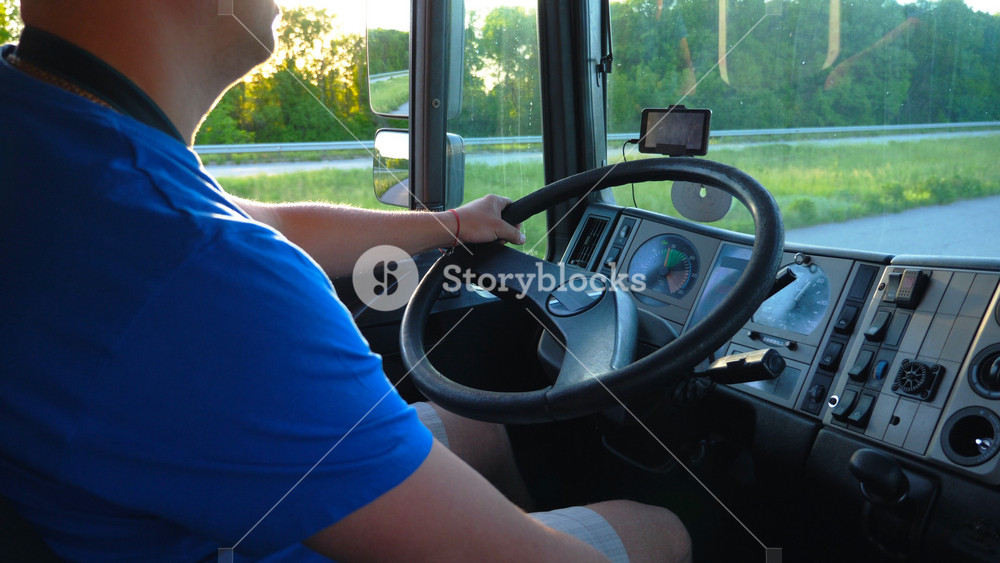 Unrecognizable man sitting behind wheel of his truck and riding through countryside. Bright sun shining at background. Close up steering wheel and dashboard of truck. View inside lorry cab. Slow mo